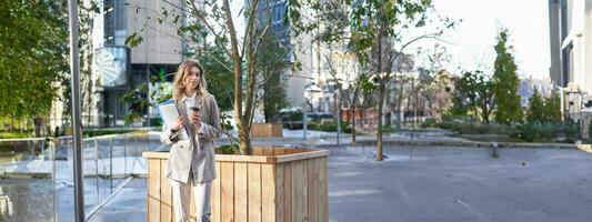 eccitato donna d'affari bevande caffè, prende porta via su pranzo vicino ufficio costruzione, in piedi su strada foto