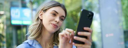 femmina dipendente seduta all'aperto e guardare a mobile Telefono, sorridente a smartphone schermo foto