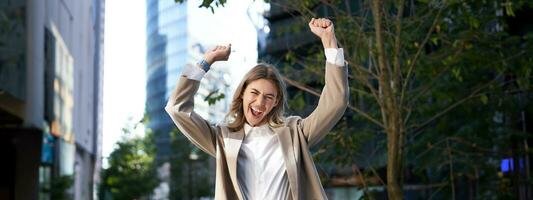 eccitato donna d'affari urla e festeggia, ascensori mani su, fa evviva gesto, celebra vittoria, raggiungere obiettivo, sta su strada foto