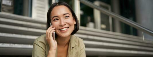 ritratto di asiatico ragazza sorrisi mentre parla su mobile Telefono. giovane donna chiamata un' amico, seduta su le scale foto