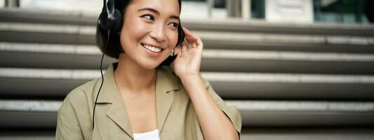 ritratto di bellissimo ragazza nel auricolari, ascoltando musica, sorridente e a piedi nel città centro foto