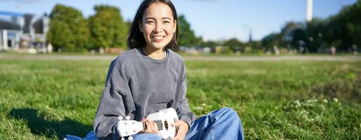 ritratto di asiatico ragazza alunno, giocando ukulele e cantando nel parco, seduta solo su coperta e godendo fabbricazione musica foto