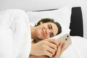 vicino su ritratto di bello giovane donna nel letto, dire bugie e andando per dormire, controllo sua Telefono messaggi, utilizzando smartphone e sorridente foto