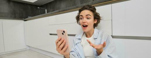 ritratto di stupito, Candido giovane donna, Tenere smartphone, reagisce sorpreso a grande notizia, video chat con qualcuno, si siede su cucina pavimento foto