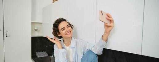 bellissimo donna prende autoscatto su smartphone, in posa nel cucina e sorridente a mobile telecamera per foto