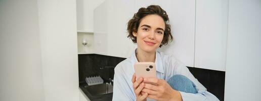 ritratto di sorridente bellissimo donna, seduta nel cucina con smartphone, guardare contento a telecamera, la spesa tempo a casa, utilizzando mobile Telefono App foto