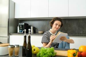 ritratto di giovane donna cucinando, scrittura Appunti, drogheria elenco nel taccuino, la creazione di elenco di pasti per cucinare attraverso pasto, seduta nel cucina vicino verdure e chopping tavola foto