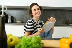 ritratto di giovane donna cucinando, scrittura Appunti, drogheria elenco nel taccuino, la creazione di elenco di pasti per cucinare attraverso pasto, seduta nel cucina vicino verdure e chopping tavola foto