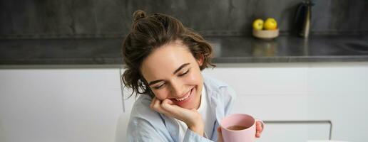 contento le mattine. ritratto di contento brunetta donna, bevande tazza di caffè nel sua cucina e sorridente, accogliente e caldo inizio di il giorno con cuppa foto