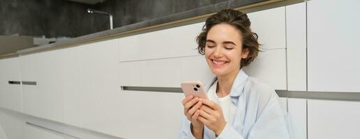 ritratto di carino sorridente donna, seduta su pavimento a casa con attraversato gambe, sembra a mobile Telefono, chat su smartphone app, orologi video nel applicazione foto