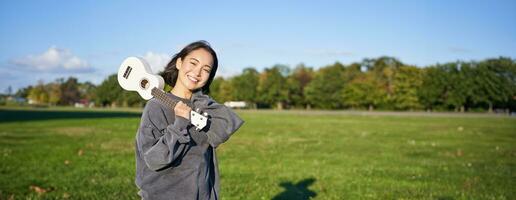 ritratto di bellissimo sorridente ragazza con ukulele, asiatico donna con musicale strumento in posa all'aperto nel verde parco foto