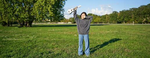 contento asiatico ragazza, musicista con ukulele, sensazione spensierato, godendo la libertà e fresco aria all'aperto, giocando musicale strumento foto