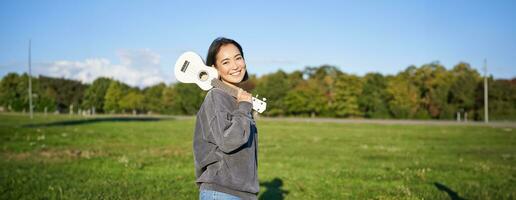 giovane fricchettone ragazza, viaggiatore Tenere sua ukulele, giocando all'aperto nel parco e sorridente foto