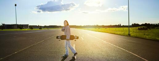 giovane pattinatore ragazza, adolescente pattinando su incrociatore, Tenere longboard e a piedi su calcestruzzo vuoto strada foto