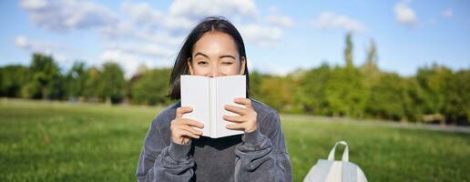 bellissimo asiatico ragazza seduta nel parco su erba, lettura e sorridente. donna con libro godendo soleggiato giorno all'aperto foto