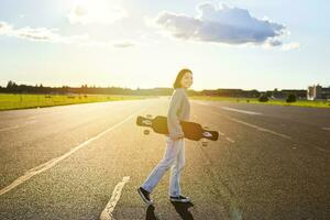 giovane pattinatore ragazza, adolescente pattinando su incrociatore, Tenere longboard e a piedi su calcestruzzo vuoto strada foto