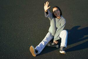 positivo coreano ragazza coperture sua viso a partire dal luce del sole, si siede su skateboard e sorrisi felicemente foto