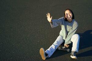 positivo coreano ragazza coperture sua viso a partire dal luce del sole, si siede su skateboard e sorrisi felicemente foto