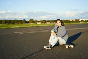 ritratto di giovane coreano ragazza seduta su sua skateboard su strada, guardare a smartphone, chat su mobile App foto