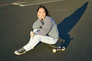 ritratto di asiatico donna seduta su skateboard, pattinando su sua incrociatore longboard, utilizzando smartphone App foto