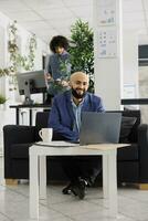 sorridente riuscito arabo uomo d'affari Lavorando su il computer portatile nel coworking spazio ritratto. contento fiducioso unicorno attività commerciale imprenditore seduta su divano a ufficio posto di lavoro e guardare a telecamera foto