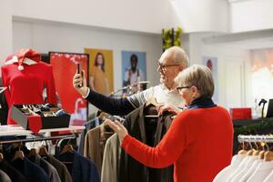 anziano coppia assunzione fotografie nel negozio, confrontando giacche su grucce con prodotti in linea su sito web. anziano persone shopping per formale Abiti durante Natale stagione, guardare per festivo vestiario.