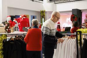 anziano persone ricerca per giacche nel shopping centro commerciale, guardare a capi di abbigliamento elementi su grucce durante inverno i saldi stagione. coppia controllo nuovo Abiti nel Al dettaglio negozio, acquisto i regali per famiglia. foto