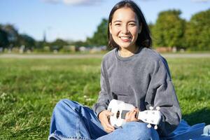 ritratto di asiatico ragazza alunno, giocando ukulele e cantando nel parco, seduta solo su coperta e godendo fabbricazione musica foto