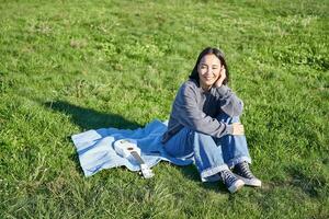 sorridente carino ragazza seduta su coperta nel parco, giocando ukulele all'aperto, godendo soleggiato caldo tempo atmosferico, indossare maglione e jeans foto