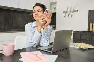 ritratto di brunetta donna chat su mobile Telefono, usi smartphone App a partire dal casa, ordini consegna mentre si siede nel cucina foto