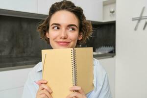 ritratto di sorridente brunetta ragazza, detiene sua pianificatore, scrive nel taccuino e sembra felice, ride, fa compiti a casa, fa Appunti, si siede a casa nel cucina foto