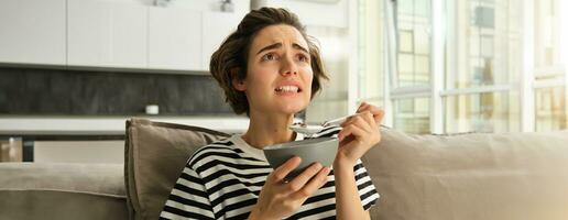 Immagine di donna mangiare prima colazione nel davanti di tv, guardare a schermo con preoccupato viso, Tenere ciotola di cereali con latte e un' cucchiaio, la spesa tempo nel vivente camera foto