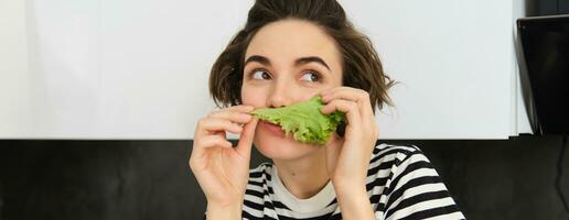 vicino su di divertente carino donna, vegetariano mangiare lattuga foglia e sorridente, concetto di salutare dieta, ragazza piace la verdura, in piedi nel il cucina foto