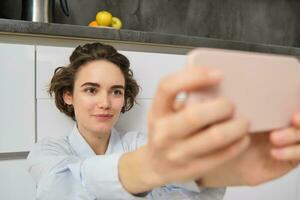 vicino su ritratto di Candido giovane donna, prende autoscatto su smartphone, utilizzando mobile Telefono foto App con filtri, si siede su cucina pavimento a casa