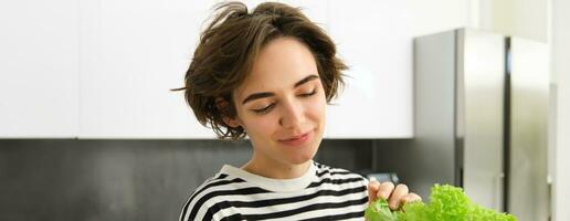 vicino su ritratto di sorridente, bellissimo femmina modello, fabbricazione un' insalata, Tenere verde lattuga foglia, preparazione la verdura, cucinando nel il cucina, in piedi nel a strisce maglietta foto