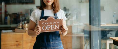 giovane femmina sospeso un' benvenuto cartello nel davanti di un' caffè negozio. bellissimo cameriera o hostess Tenere un' tavoletta preparazione nel un' ristorante. foto