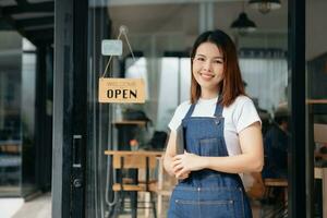 giovane femmina sospeso un' benvenuto cartello nel davanti di un' caffè negozio. bellissimo cameriera o hostess Tenere un' tavoletta preparazione nel un' ristorante. foto