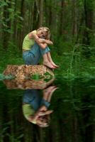 adolescenziale ragazza si siede su un' ceppo nel un' naturale foresta parco con un' triste e riflessivo Guarda, riflessa nel il acqua foto