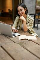 ritratto di elegante urbano donna Lavorando su il computer portatile a partire dal all'aperto bar, seduta con documenti e computer, sorridente foto