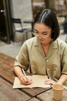 verticale tiro di giovane asiatico donna fare compiti a casa, fabbricazione Appunti, scrittura qualcosa fuori uso, seduta nel un all'aperto bar e potabile caffè foto