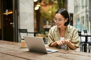 giovane donna seduta su in linea incontro nel all'aperto bar, parlando per il computer portatile telecamera, spiegando qualche cosa, potabile caffè foto