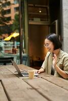 ritratto di asiatico donna guardare a computer portatile, video Chiacchierare, parlando con qualcuno attraverso computer telecamera, seduta nel bar e potabile caffè, in linea incontro foto