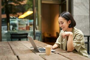 ritratto di asiatico donna guardare a computer portatile, video Chiacchierare, parlando con qualcuno attraverso computer telecamera, seduta nel bar e potabile caffè, in linea incontro foto