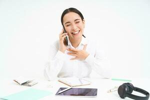 Immagine di asiatico donna nel ufficio, parla su cellulare, discutere opera con cliente su mobile Telefono, si siede al di sopra di bianca sfondo foto