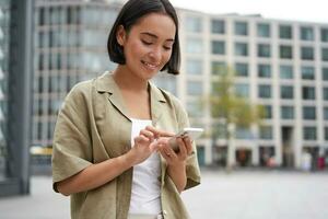 mobile tecnologia. sorridente asiatico donna utilizzando smartphone app, guardare a sua telefono su strada, controllo carta geografica, chiamata o sms qualcuno foto