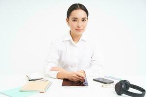 sorridente asiatico donna si siede nel sua ufficio, lavori nel digitale tavoletta, ha cuffia, smartphone e opera documenti su tavoletta, bianca sfondo foto