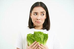 salutare dieta e crudo cibo. triste e infelice asiatico donna, Tenere fresco lattuga foglie, cavolo nel mani, mangiare la verdura, bianca sfondo foto