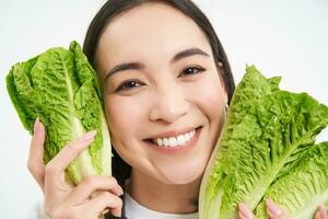 vicino su ritratto di contento coreano donna, Spettacoli sua viso con lattuga, mangiare cavolo, piace la verdura, si sente salutare e energizzato, bianca sfondo foto