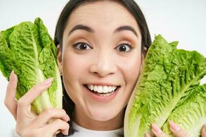 vicino su ritratto di contento coreano donna, Spettacoli sua viso con lattuga, mangiare cavolo, piace la verdura, si sente salutare e energizzato, bianca sfondo foto