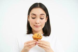 vicino su ritratto di asiatico ragazza con delizioso cupcake, Guarda pensieroso a dolce mentre su dieta, controllo calorie, bianca sfondo foto